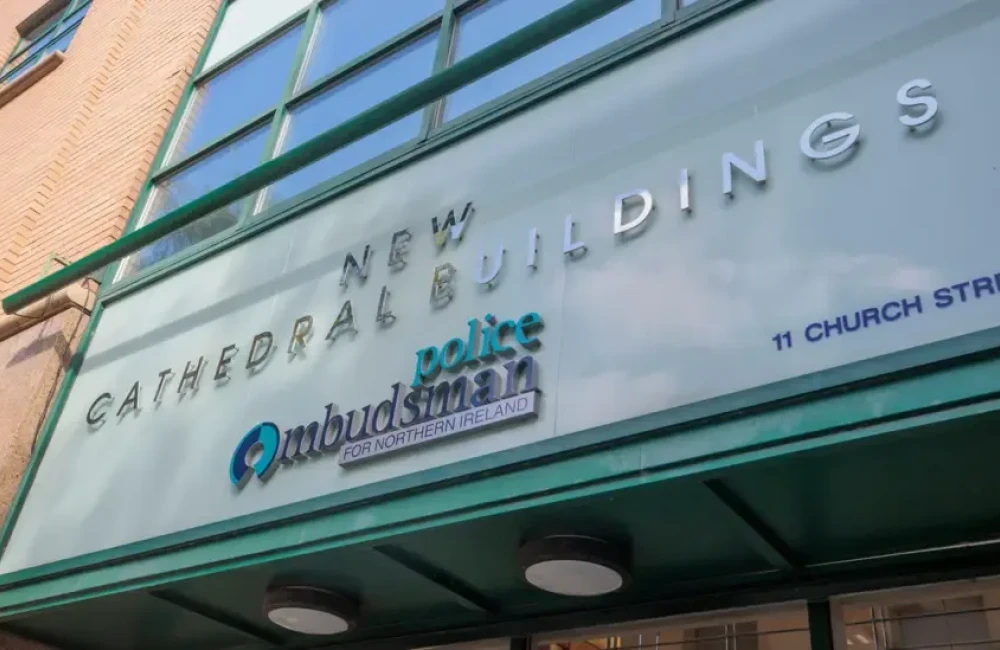 Above the entrance to the police ombudsman's office a sign reads 'NEW CATHEDRAL BUILDINGS:  police Ombudsman FOR NORTHERN IRELAND'. The lettering is silver, navy and green, and is set against a background of lighter, pastel green.