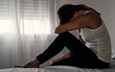 Anonymous woman sitting on bed with her head in her arms. She is wearing black trousers and a white vest. Her dark hair is tied in a ponytail.