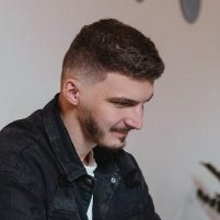 A young man smiles slightly, he appears to be seated and is looking at a screen, possibly of a laptop.