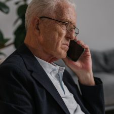 An elderly man is speaking on his mobile phone. His expression is serious and the room is slightly darkened. He is wearing a black suit jacket and light colourede open necked shirt.
