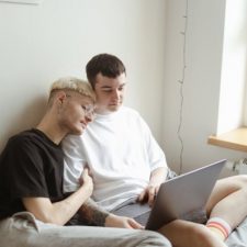 A young gay couple sit on a bed together, the man on the left is wearing a white jumper and has dark hair, he is using a laptop. The man on the right is wearing a black tshirt and grey trousers, he has blonde hair and has his head leaning on the other man's shoulder, and his hand on the man's right arm.