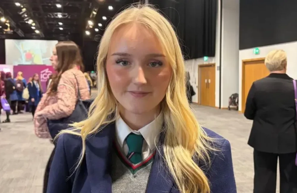 Jessica is looking at the camera. She has long blonde hair and is wearing a navy school blazer and a teal coloured tie with a grey jumper.