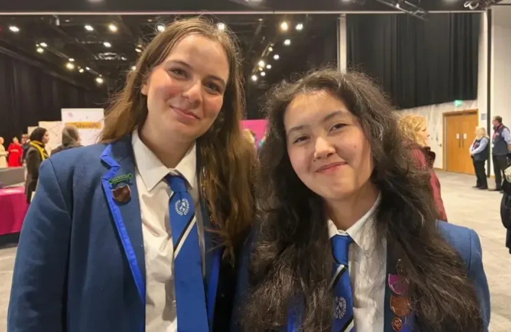 Eve and Anna are smiling at the camera. Eve has light brown hair and Anna has dark brown hair. They are wearing navy school blazers that have lighter blue lining. Their school ties are light blue to match.