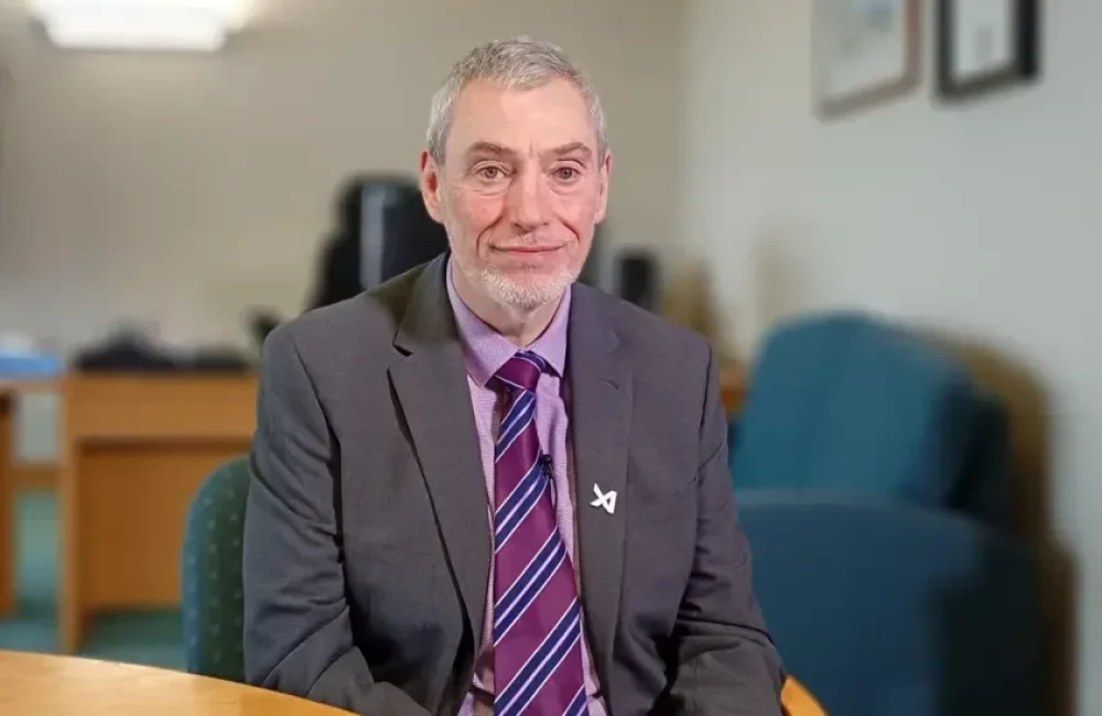 Hugh Hume. He has short grey hair, wearing a dark grey blazer with a silver sin on the lapel, light purple shirt and purple striped tie. He is sitting on a chair beside a table in an office.
