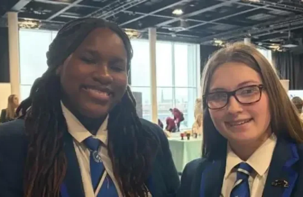 Kayla and Brooke and smiling at the camera. Brooke wears glasses. They are wearing navy school blazers that have lighter blue lining. Their school ties are light blue to match.