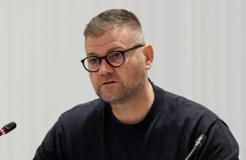 Colin is sat down in front of two small microphones. Ge is wearing a black t-shirt and dark round-rimmed glasses. His fair is greying and short and he has a short grey-speckled stumble.
