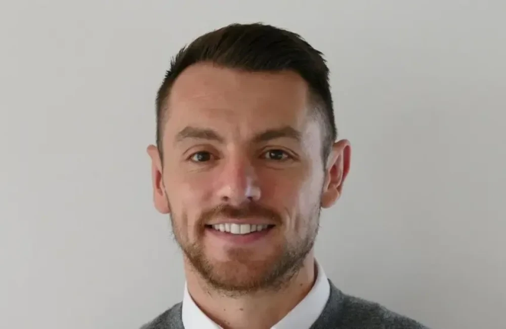 A man with dark hair smiling. He is wearing a grey jumpter and a white shirt.