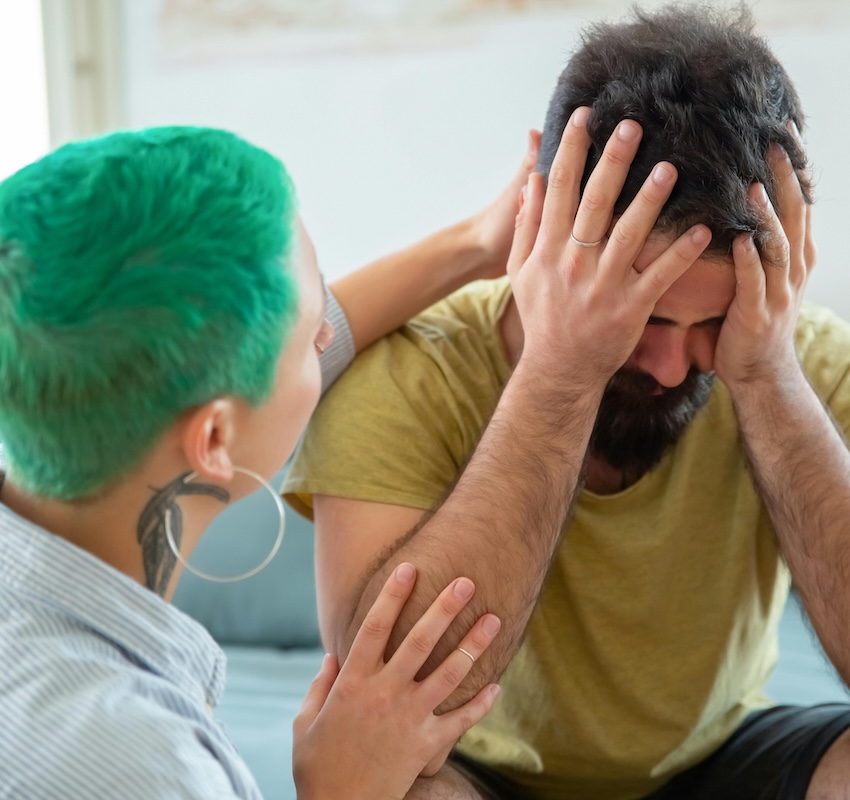 A man sits with his head in his heands, he is wearing a yellow tshirt and has short dark hair and a beard. He is being comforted by a woman who has one arm around him, she has close cropped bright green hair, large hoop earrings and a tattoo on her neck below her right ear, wearing a striped white shirt.