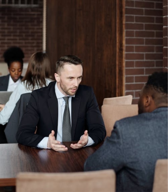 Two men dressed in suits sit together at a table in a red-brick-walled room, having a serious conversation.