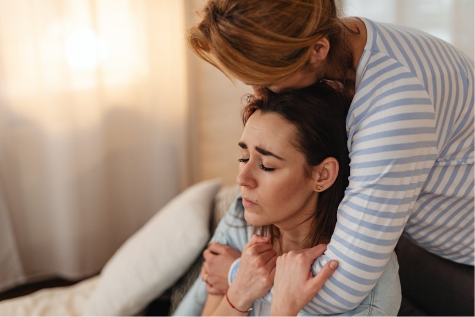 A woman stands behind a younger woman in a seated position, she has her arms around the younger woman who has her eyes closed.