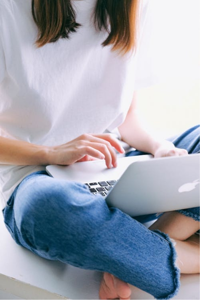 A woman in a white top and blue jeans sits crosslegged while using a laptop.
