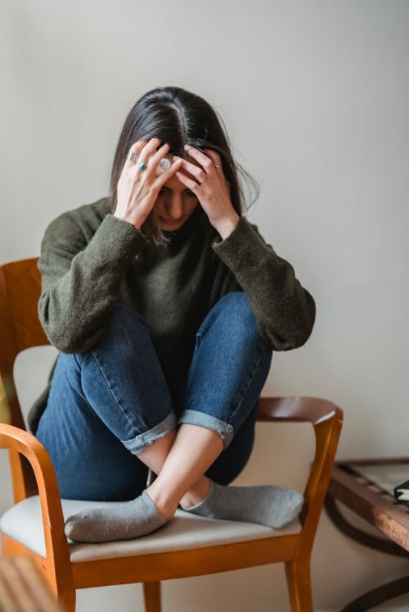 A woman sits crosslegged in a chair with her head in her hands.