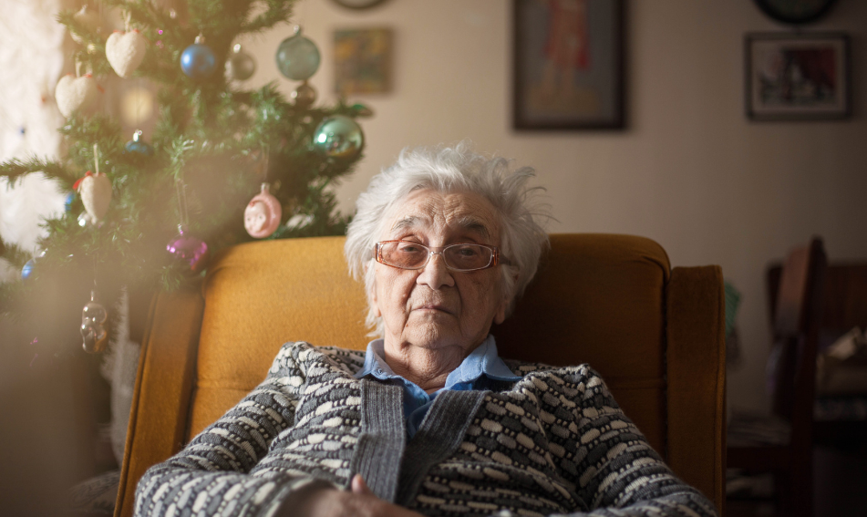 Older woman sits in front of a Christmas tree seeming sad.